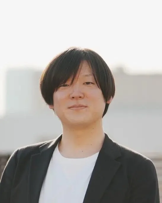 明るい屋外で撮影された、黒髪のアジア人男性のポートレート。白いTシャツと黒いジャケットを着用し、カメラにまっすぐな視線を向けている。