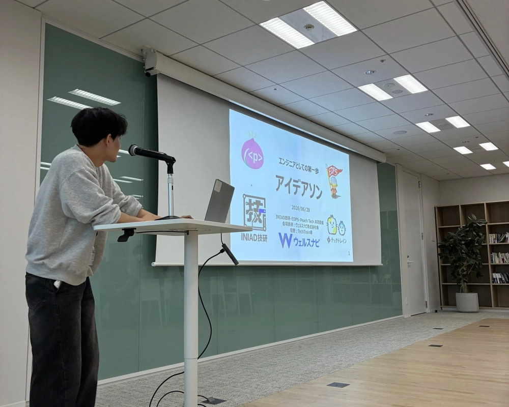 男性が演台でプレゼンテーションを行っており、スクリーンには「エンジニアとしての一歩 アイデアソン」というタイトルと日付、共同開催・協賛企業名が表示されています。
