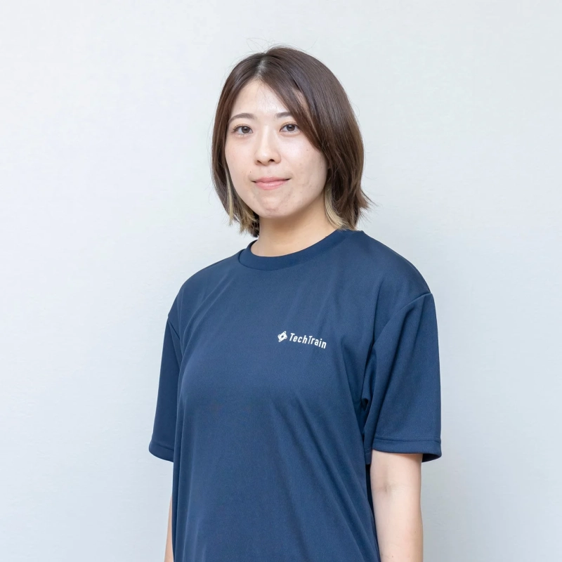 白い背景の前に立つ、TechTrainのロゴ入り紺色Tシャツを着たショートヘアの女性のポートレート。