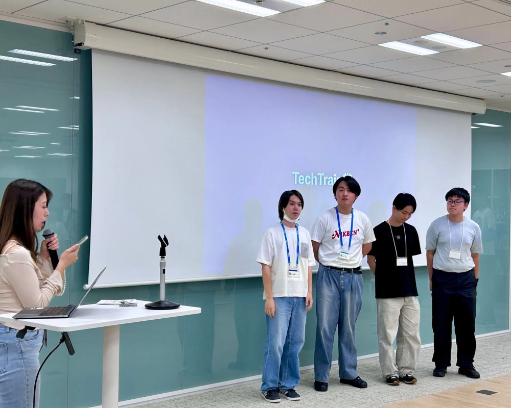 会議室で女性がマイクを持ちプレゼンテーションを行っており、スクリーンの前には4人の若い男性が立っています。