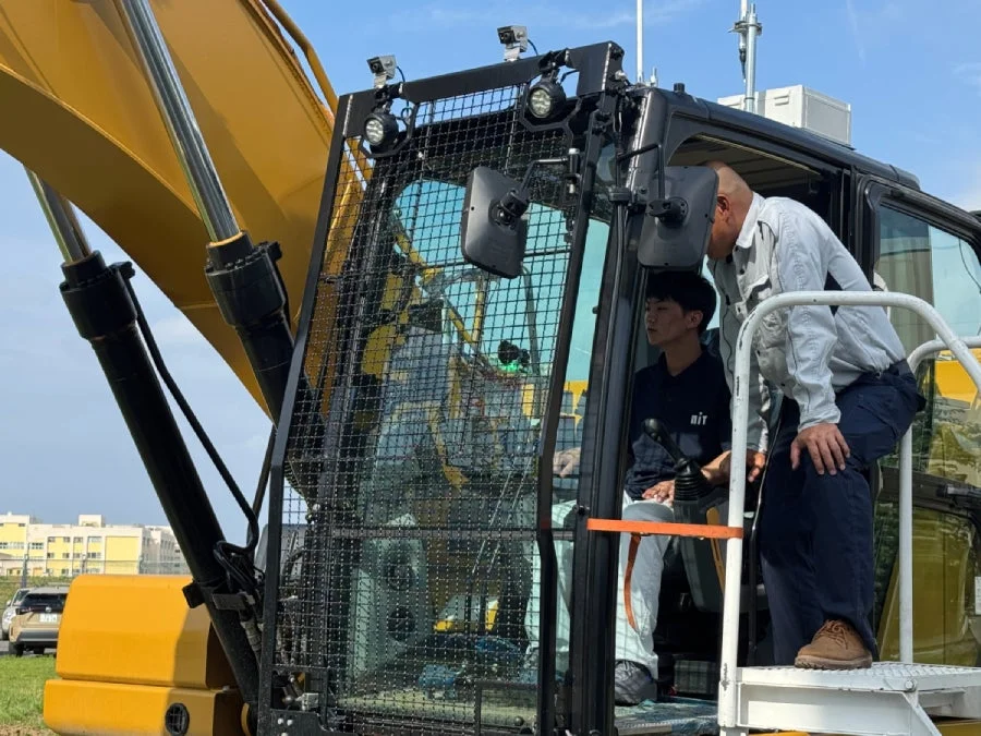 黄色の油圧ショベルの運転席に座る若い男性と、その横で指導する年配の男性が写っている様子。
