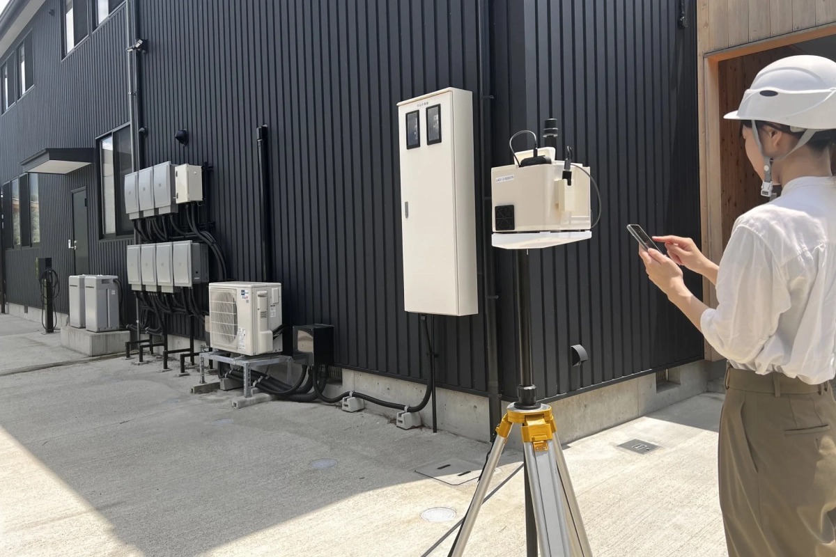 屋外でヘルメットを着用した作業員がスマートフォンを操作し、測量機器を使って建物の設備を点検している