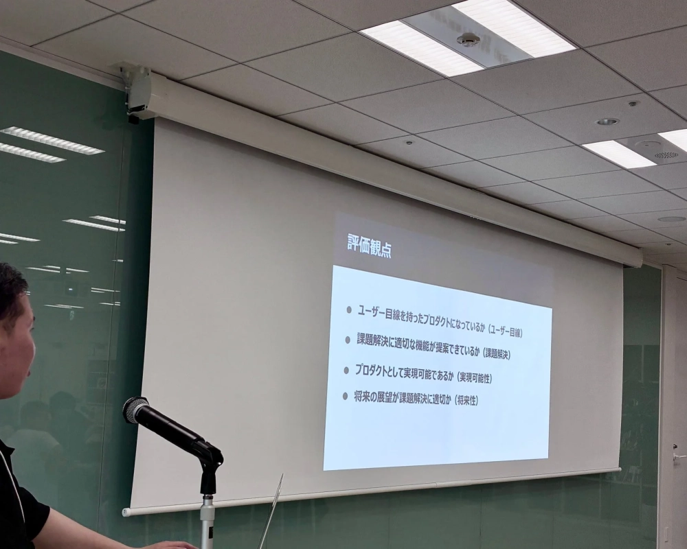 会議室でプレゼンテーションが行われており、スクリーンには「評価観点」のスライドが映し出されている。