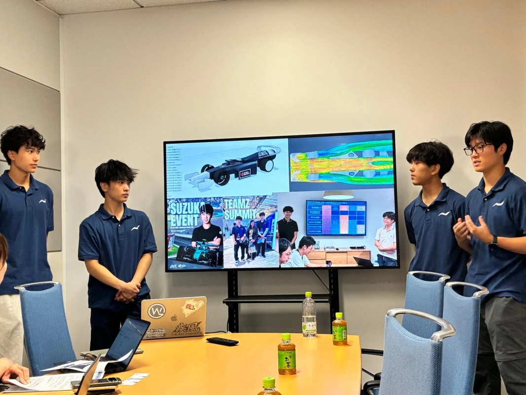 会議室で数名の若者が大型モニターを使い、レーシングカーの設計や流体解析に関するプレゼンテーションを行っています
