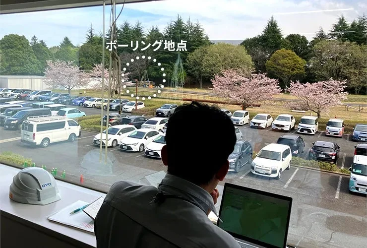 窓越しに駐車場と桜並木を眺める人物、遠方にボーリング地点
