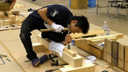 若い男性が白い手袋を着用し、ノミと木槌を使って真剣な表情で木材加工を行っている