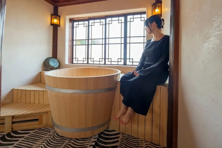 大きな木製浴槽がある和モダンな空間で、窓辺に座り外を眺める女性の姿
