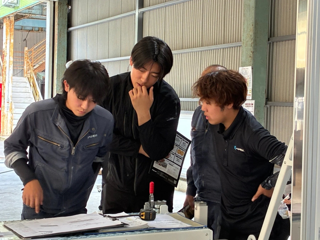 倉庫のような場所で、3人の若者が書類を囲んで真剣に打ち合わせや作業内容の確認をしている様子
