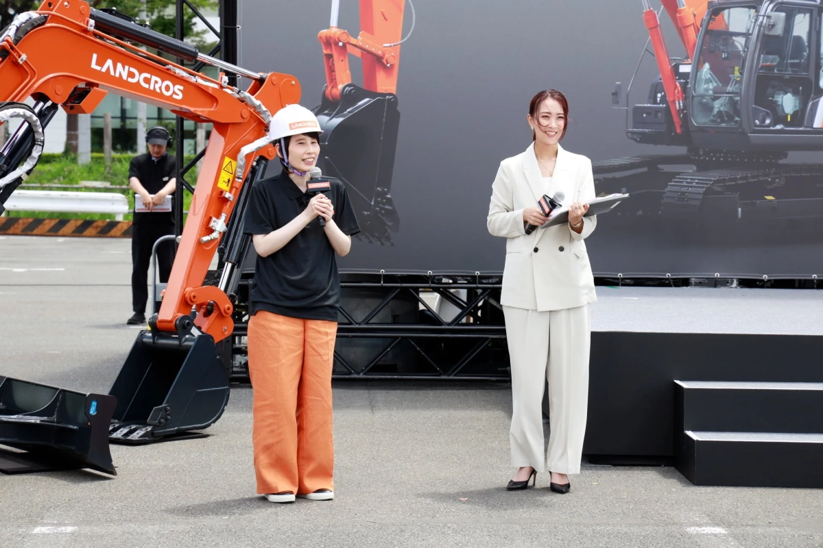 屋外のイベント会場で、オレンジ色の油圧ショベルを背景に2人の女性が立っています。一人はヘルメットを着用しマイクを持って話し、もう一人は白いスーツ姿で資料を持っています。LANDCROSというブランドの発表会のようです。
