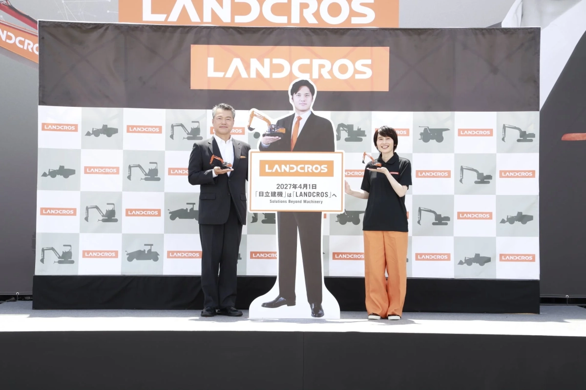 LANDCROSのロゴが掲げられたステージで、2人の人物が記念撮影をしている様子です。中央のボードには「2027年4月1日 『日立建機』は『LANDCROS』へ Solutions Beyond Machinery」と書かれており、新ブランド発表会と見られます。