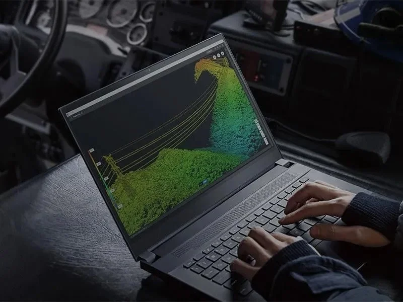 車内でノートパソコンを操作する人物。画面には電力線のような構造物を含む3Dの点群データが表示されており、現場でのデータ分析や測量作業を示唆しています。