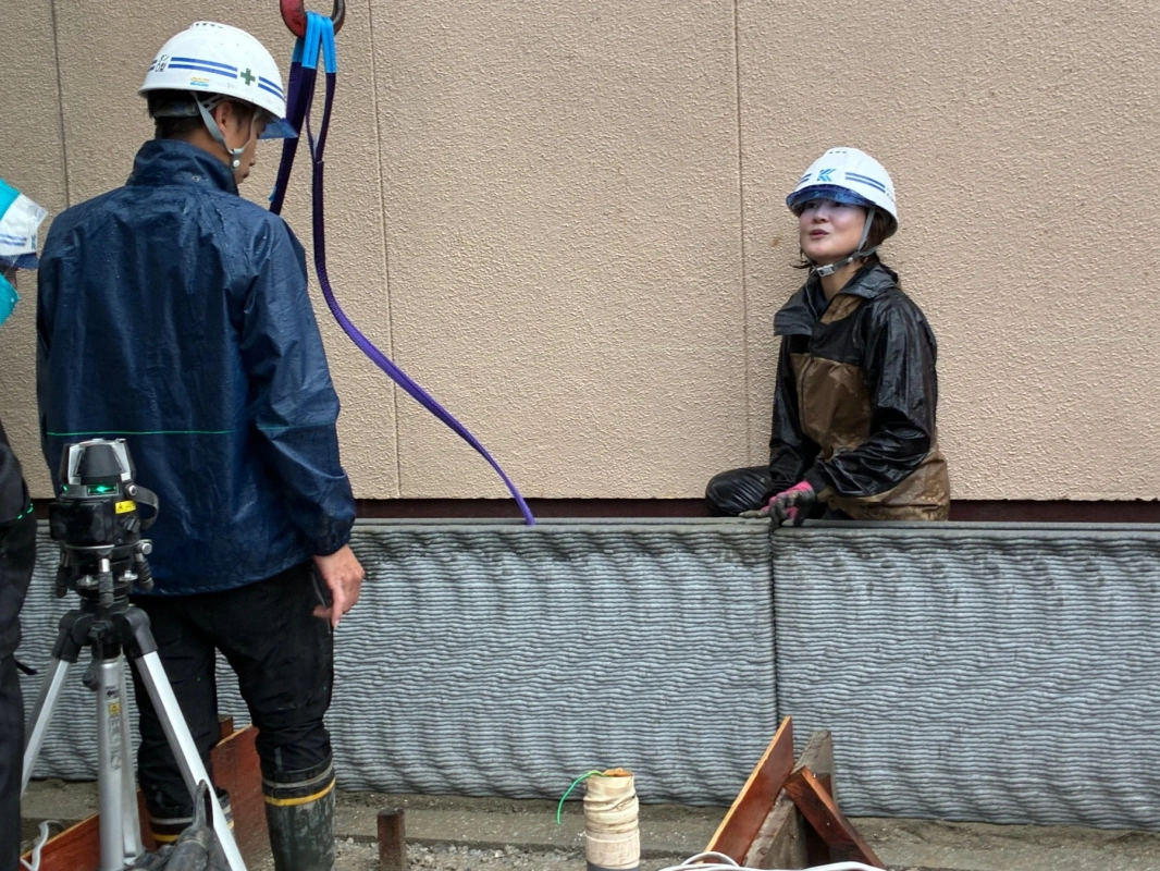 雨が降る建設現場で、ヘルメットと雨具を着用した2人の作業員が、独特のテクスチャを持つ壁の横にいる様子
