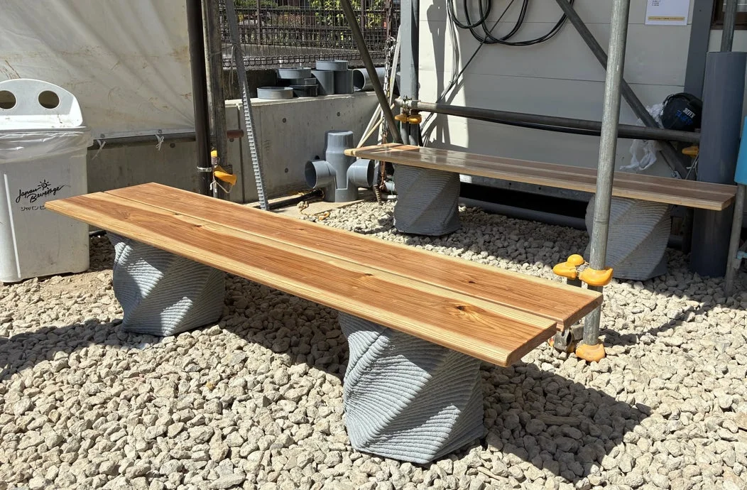 木製の座面とユニークなデザインのコンクリート製脚を持つベンチが、砂利が敷かれた屋外に設置されている様子