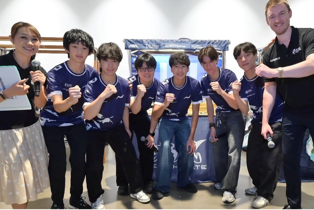 STEMレーシングイベントに参加していると思われる、お揃いの青いシャツを着た学生チームが、女性プレゼンターと男性メンターと共にガッツポーズをしている集合写真