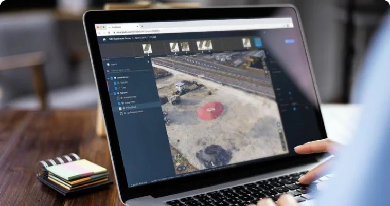 ノートパソコンで作業する人物が、ドローンで撮影された建設現場の3Dモデルや測量データを表示するソフトウェアを使用している様子